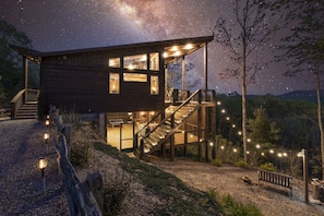 A modern wooden cabin with a staircase leading to a balcony, surrounded by trees and a starry night sky.