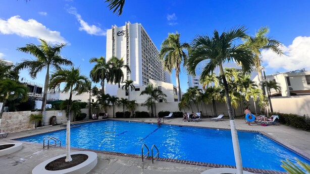 Piscina cercada por palmeiras e um edifício alto com 'es' nele.