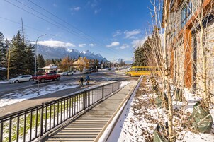 Rua coberta de neve com um ônibus amarelo, uma passarela e edifícios com montanhas ao fundo.