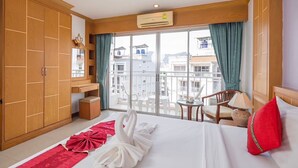 A hotel room with a large bed, a wooden wardrobe, a balcony with a view of buildings, and a small table with chairs. - Sun Shine Patong (Patong)