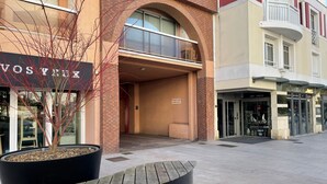 A building with a brick facade, a large black planter with a tree, and storefronts with signs like "VOS THUX" and "ART - DECO".