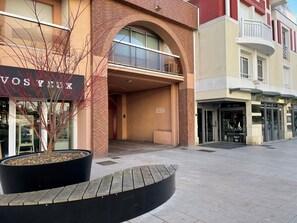 A building with a brick facade, a large black planter with a tree, and storefronts with signs like "VOS THUX" and "ART - DECO". - Résidence de la Plage by Interhome (Arcachon)