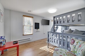 Une chambre avec un lit superposé, un téléviseur fixé au mur, une fenêtre avec des stores, une table rouge avec un sac et un téléphone, et une chaise colorée.