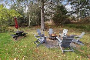 Un coin pique-nique aménagé avec un foyer et des chaises Adirondack dans une zone boisée.