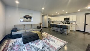 Ein modernes Wohnzimmer mit grauem Sofa, einem gemusterten Teppich und einer Küchenzeile mit weißen Schränken und einem Edelstahlkühlschrank.