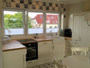 A kitchen with white cabinets, a stove, and a sink. There are two windows with a view of buildings and trees.