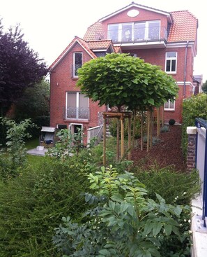 A two-story brick house with a balcony, a wooden pergola, and a garden with various plants.