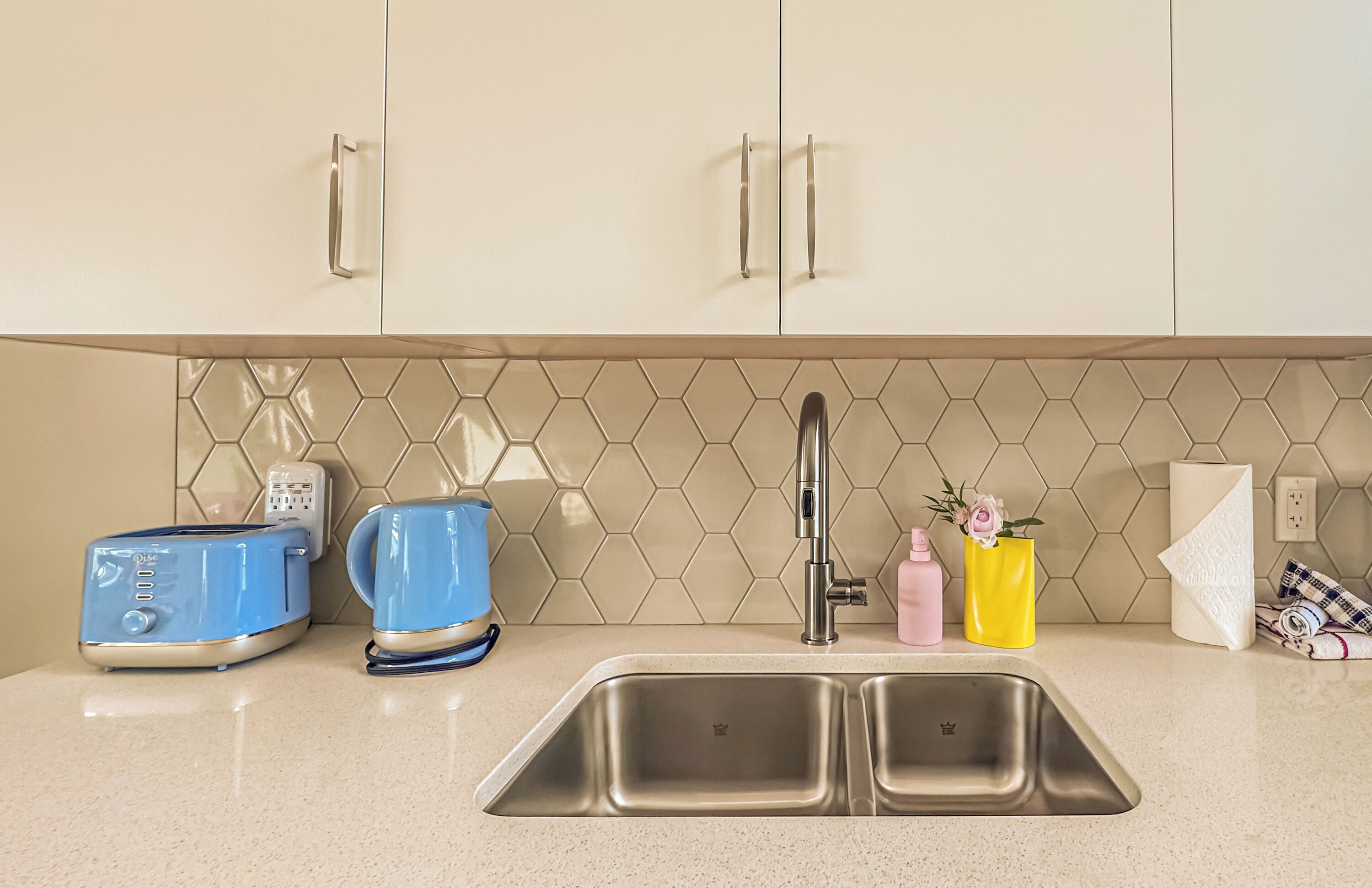 A kitchen with a stainless steel sink, two blue toasters, a coffee maker, and a hexagonal tile backsplash.