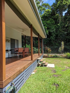 Un espacio al aire libre cubierto con una terraza de madera, una mesa y sillas, y vistas a un jardín exuberante con árboles y una valla.