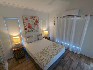 Un dormitorio con cama, dos mesitas de noche, aire acondicionado de pared y ventana con cortinas.