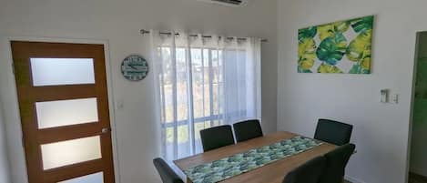 A dining area with a wooden table, black chairs, a patterned table runner, a clock, and a window with sheer curtains.