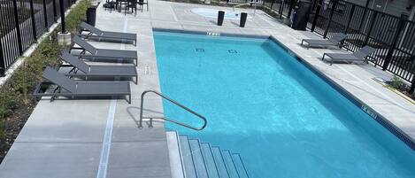 A rectangular swimming pool with a metal ladder, surrounded by a black metal fence. There are lounge chairs and tables on the poolside.