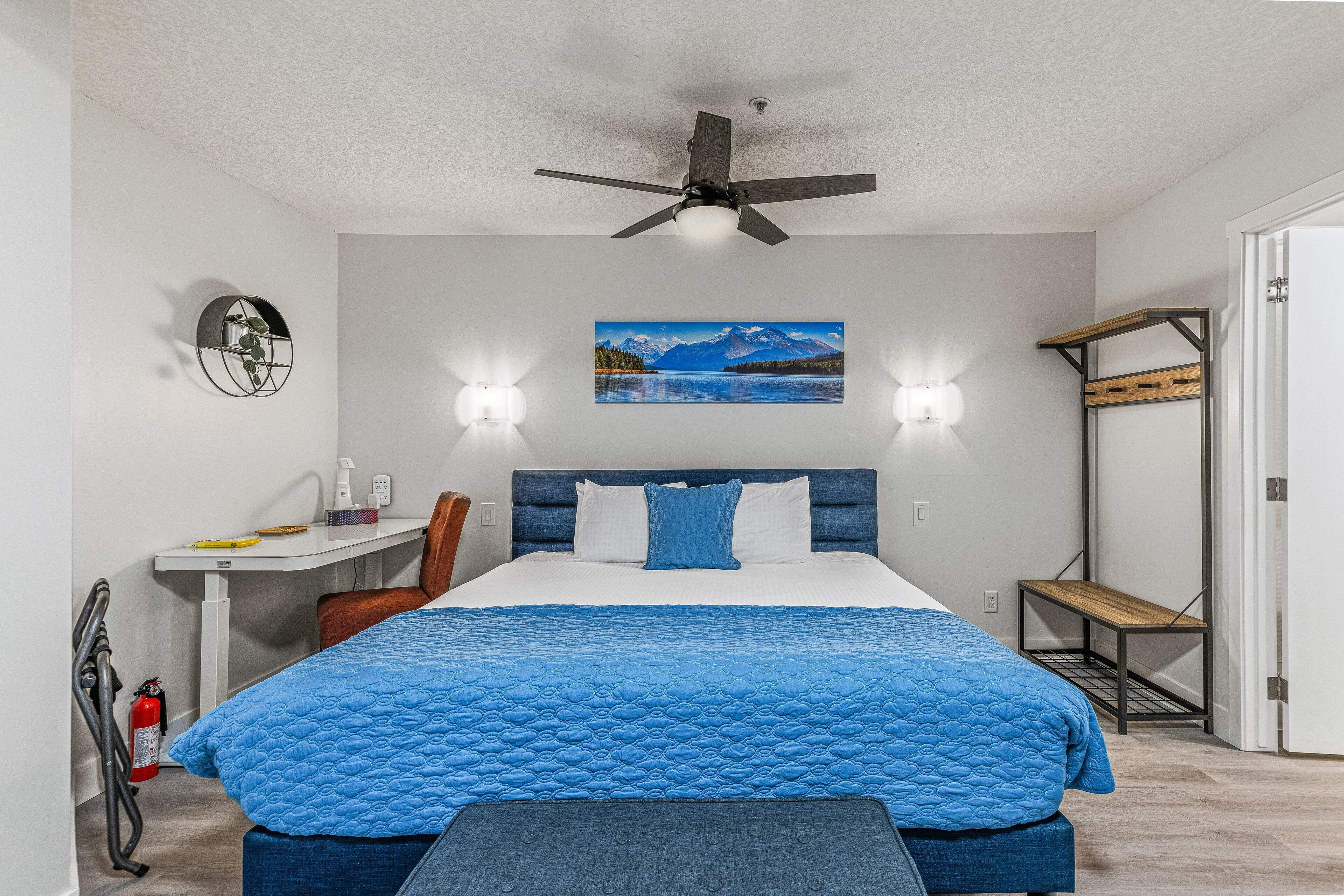 A bedroom with a bed, a desk, a chair, a ceiling fan, a wall-mounted shelf, and a door.