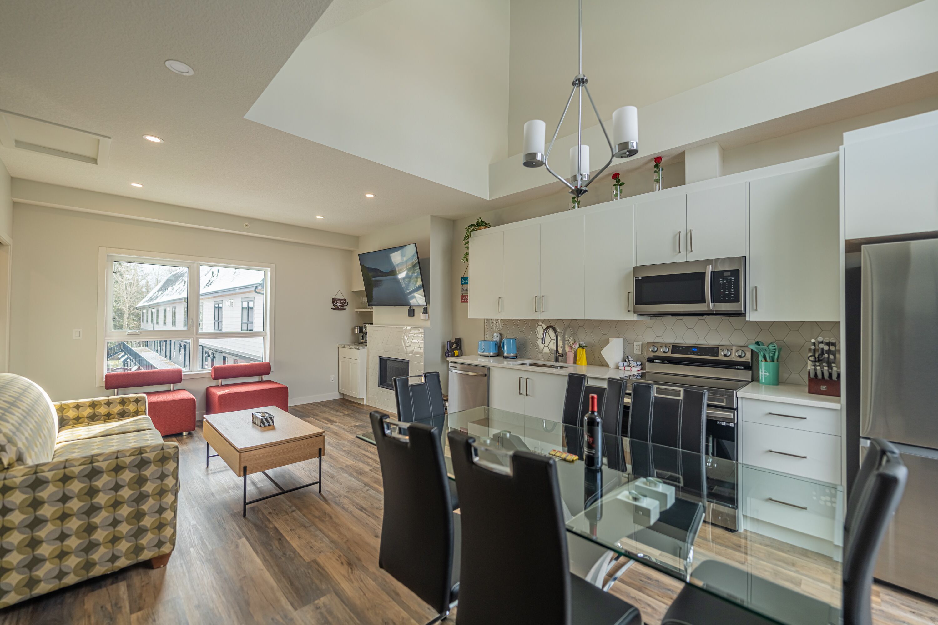 A modern kitchen with white cabinets, a microwave, and a refrigerator. There is a dining table with chairs and a sofa in the living area.