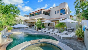Un área de piscina con sillones de descanso y sombrillas, una zona de barbacoa y una casa moderna con piscina.