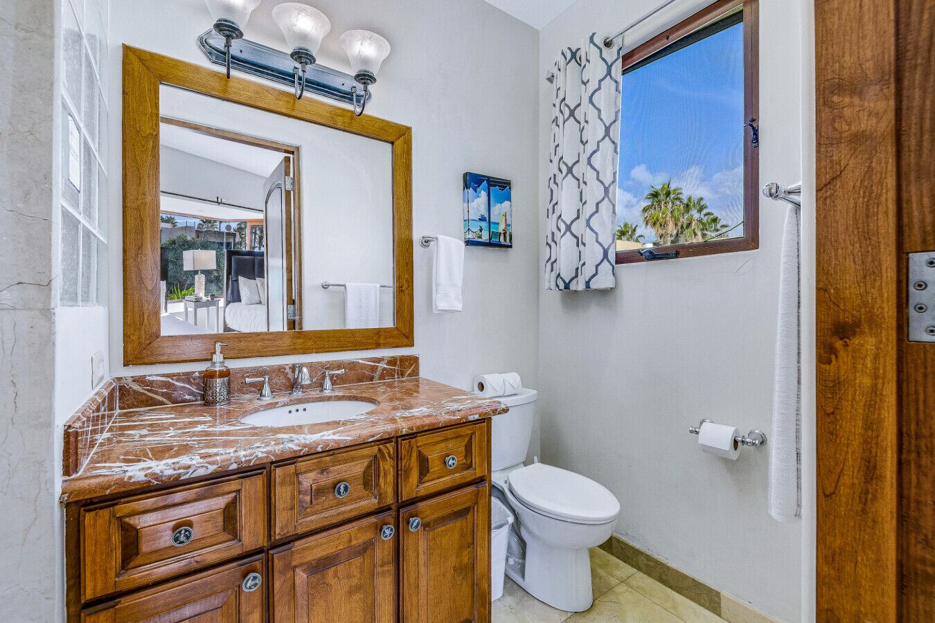 A bathroom with a marble sink, wooden vanity, a large mirror, a toilet, and a window with a view of palm trees.