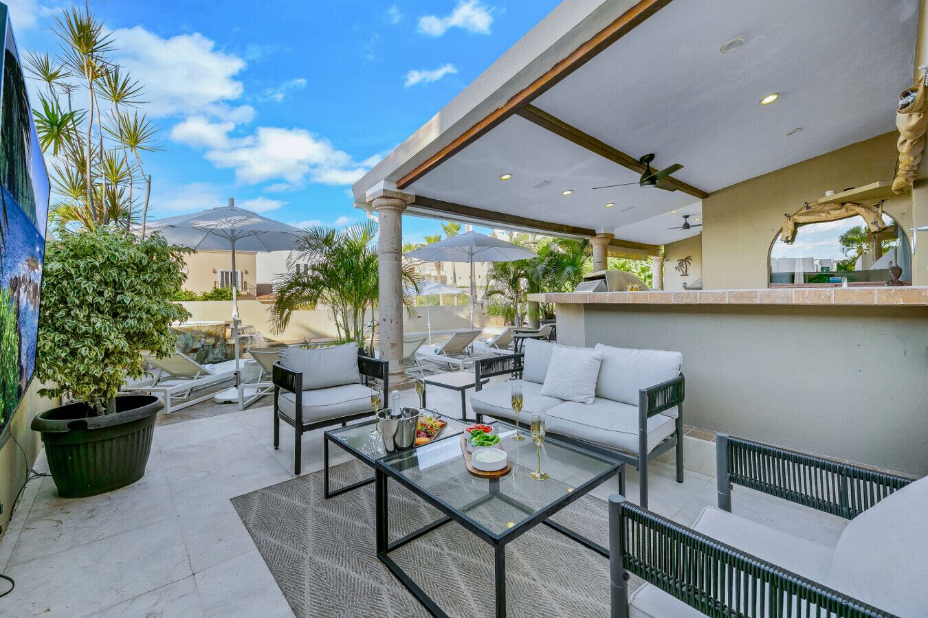 Area patio esterna con tavolino in vetro, divani e poltrone bianchi. Sono presenti ombrelloni, una pianta in vaso grande e vista sugli edifici circostanti.