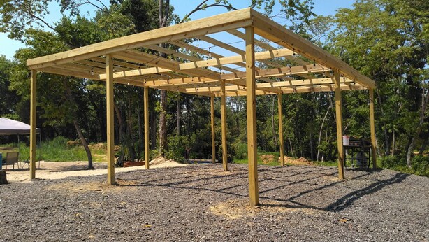 Una estructura de pérgola de madera con base de grava, rodeada de árboles y una tienda de campaña al fondo.