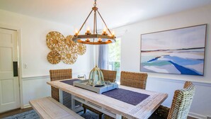 A dining area with a wooden table, wicker chairs, a bench, a chandelier, and a large abstract painting on the wall. - Relax in Renovated Home w/ Private Plunge Pool; Screened Porch; 2 Master Suites; 4 Min Walk to Beach (Isle of Palms)