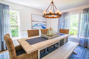 A dining area with a wooden table, wicker chairs, a chandelier, and a view of greenery through large windows.