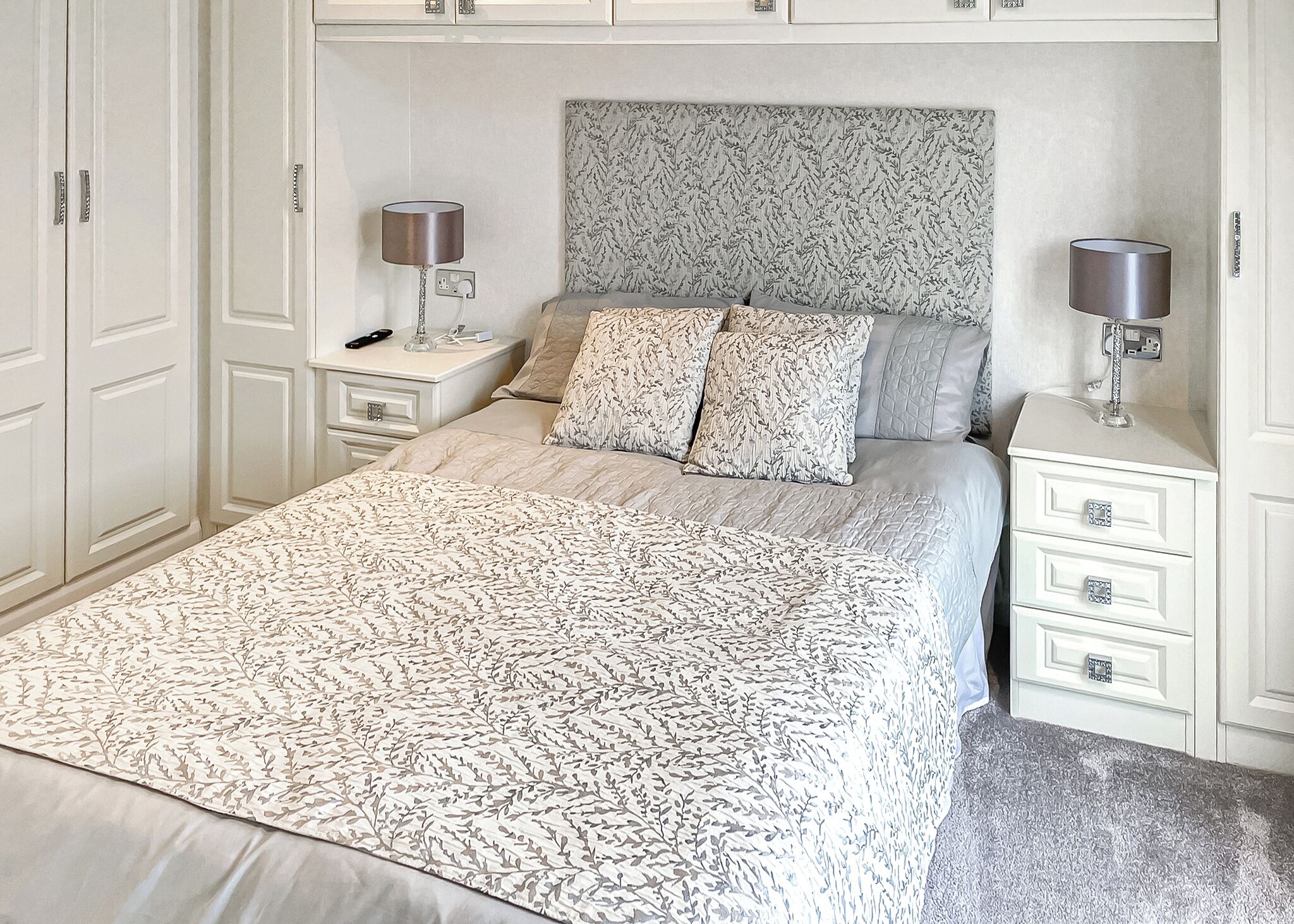 A bedroom with a large bed, two bedside tables, a headboard with a leaf pattern, and a carpeted floor.