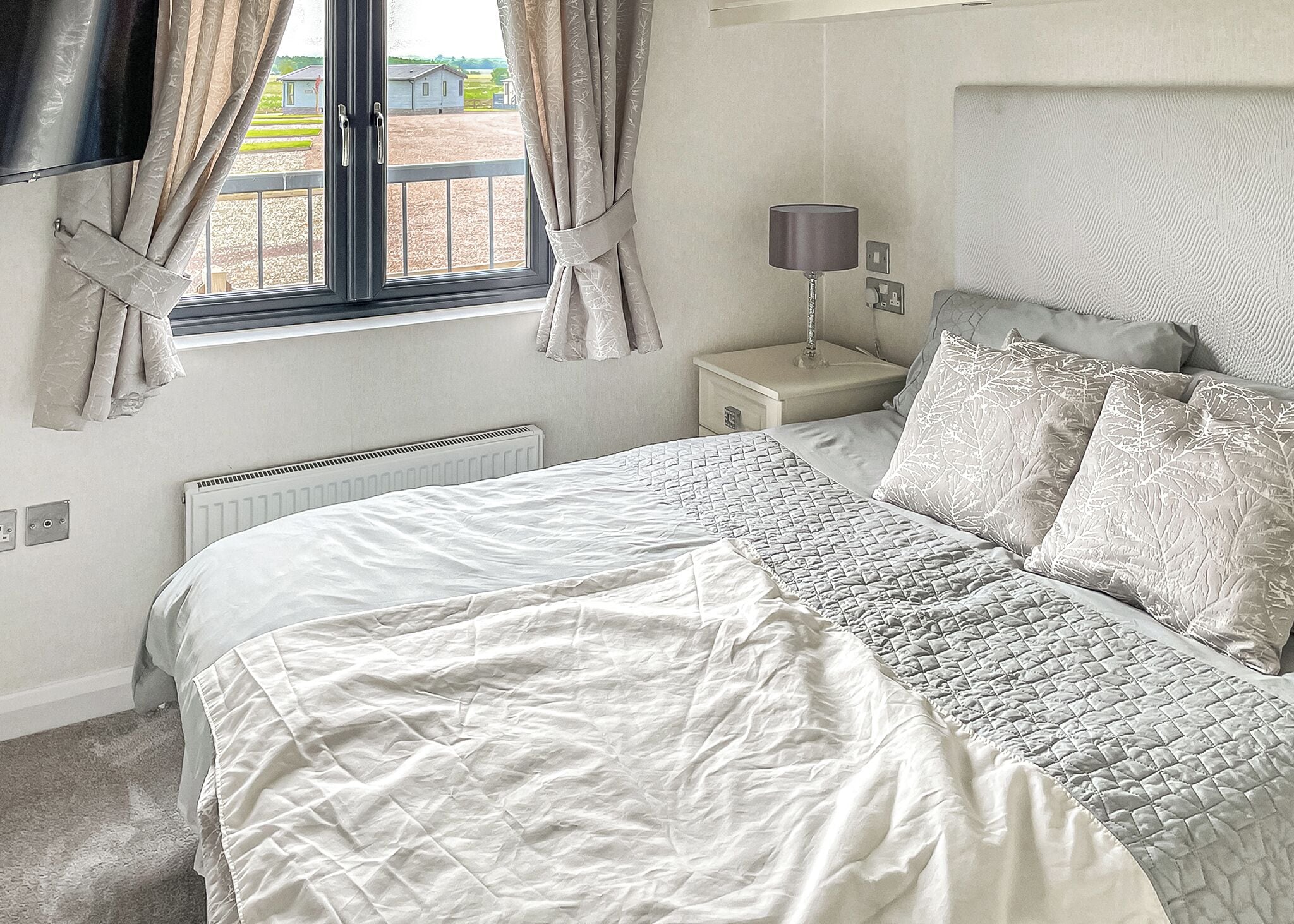 A bedroom with a large bed, a bedside table with a lamp, a window with curtains, and a radiator.
