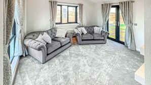 A living room with two grey sofas, a patterned carpet, and large windows with curtains.