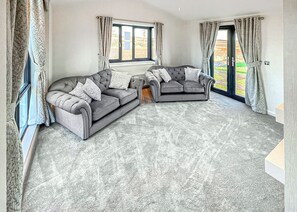 A living room with two grey sofas, a patterned carpet, and large windows with curtains.