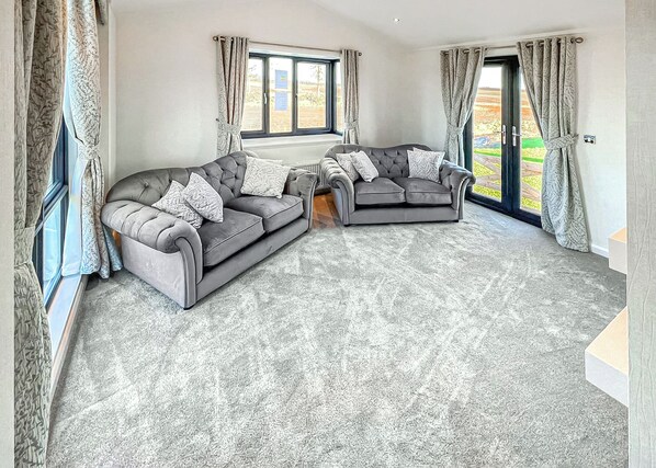 Un salon avec deux canapés gris, une moquette à motifs et de grandes fenêtres avec des rideaux.