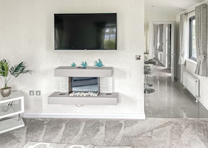 A modern living room with a flat-screen TV mounted above a fireplace, a potted plant, and a hallway leading to another room.
