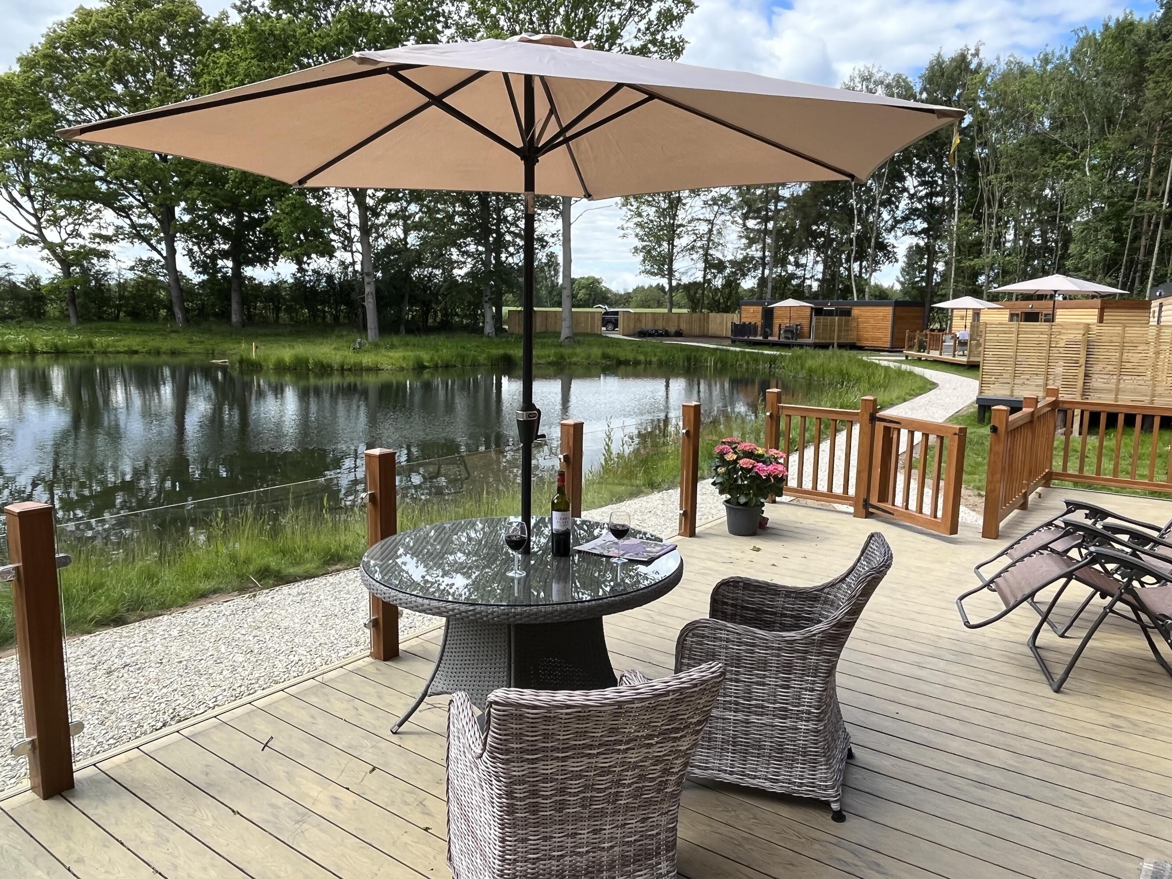 A deck with a round table and wicker chairs, a large umbrella, and a view of a lake.