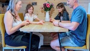 Une famille de quatre personnes est assise autour d’une table en bois, devant un vase blanc rempli de fleurs, dans une pièce aux murs en bois et où se trouve une chaise jaune.