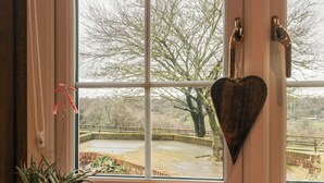 Una ventana con un adorno en forma de corazón, una planta en maceta y vistas de un parque con un muro de ladrillo y árboles.