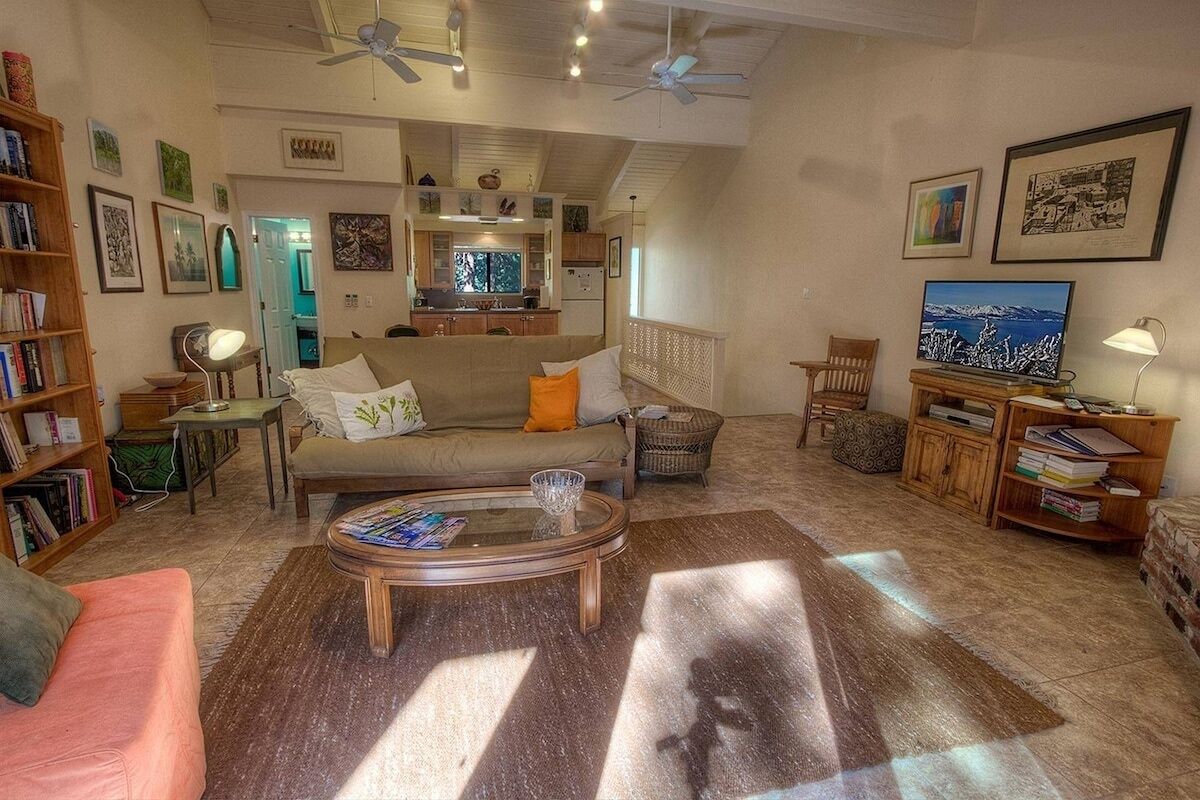 A spacious living room with a sofa, coffee table, and a television. There is a bookshelf and a dining area in the background.