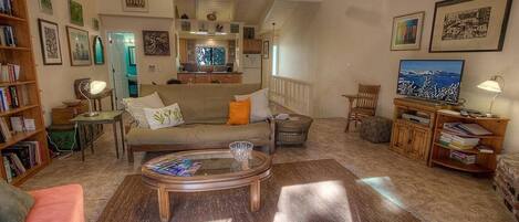 A spacious living room with a sofa, coffee table, and a television. There is a bookshelf and a dining area in the background.
