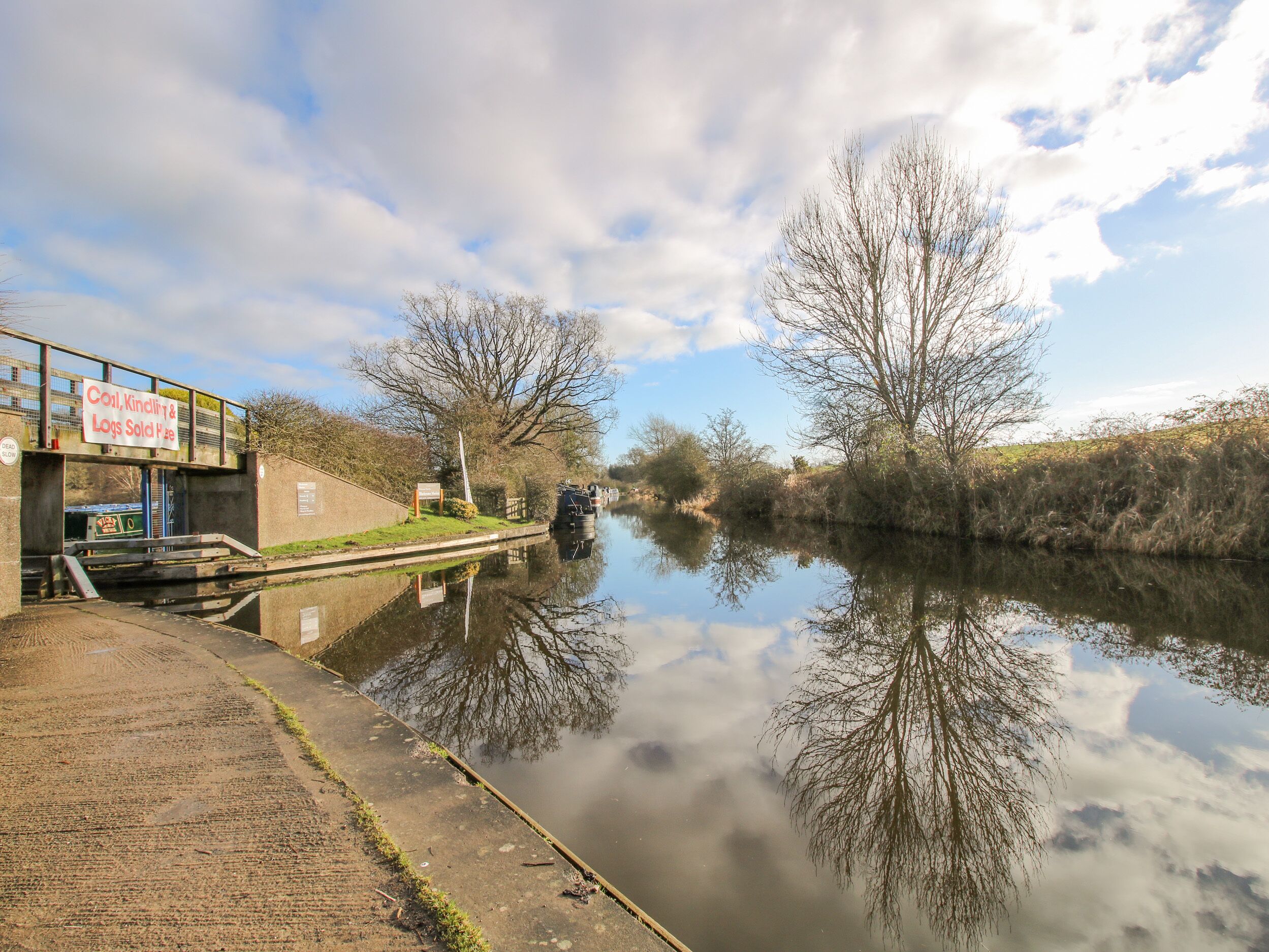 A canal with a lock gate, a signboard advertising coal, kindling, and logs, and a clear sky.