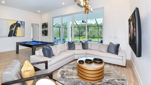 A modern living room with a sectional sofa, a pool table, and a large window offering a view of the outdoors. - Victor's Villa Score a Winning Vacay: Superhero BR, Theater, More, Near Parks (Davenport)