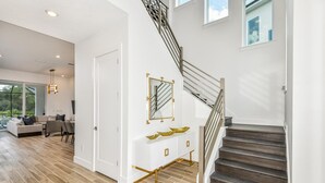 A modern interior with a staircase, a white console table with gold accents, and a large window overlooking a garden. - Victor's Villa Score a Winning Vacay: Superhero BR, Theater, More, Near Parks (Davenport)