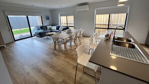 Eine moderne Küche mit Kochinsel, ein Essbereich mit weißen Stühlen sowie ein Wohnzimmer mit dunklem Sofa und grünen Kissen.