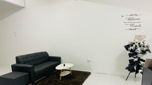 Ein modernes Wohnzimmer mit einem dunkelgrauen Sofa, einem kleinen weißen Tisch und einem dunklen Teppich. Ein dekoratives Wandschild zeigt den Schriftzug 