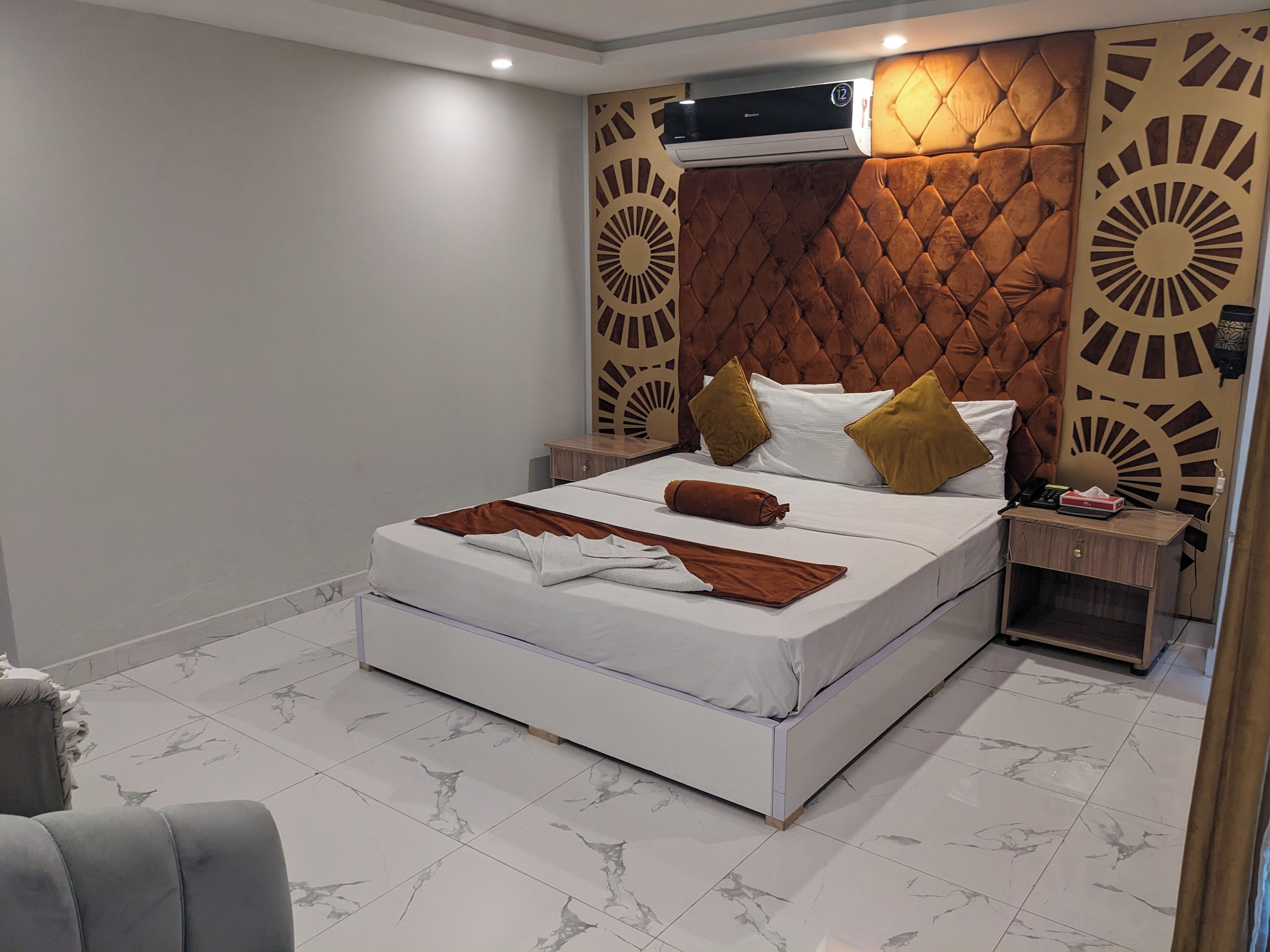 A modern bedroom with a tufted headboard, a white bed with brown and yellow pillows, a wooden nightstand, and a marble-tiled floor.