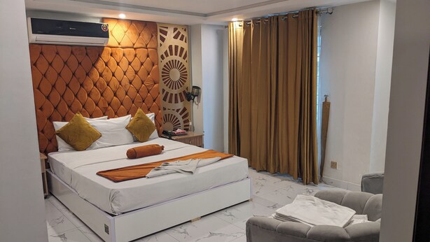Un dormitorio moderno con un cabecero capitonado, una cama grande, una mesita de noche y un sillón gris, junto a una ventana con cortinas.