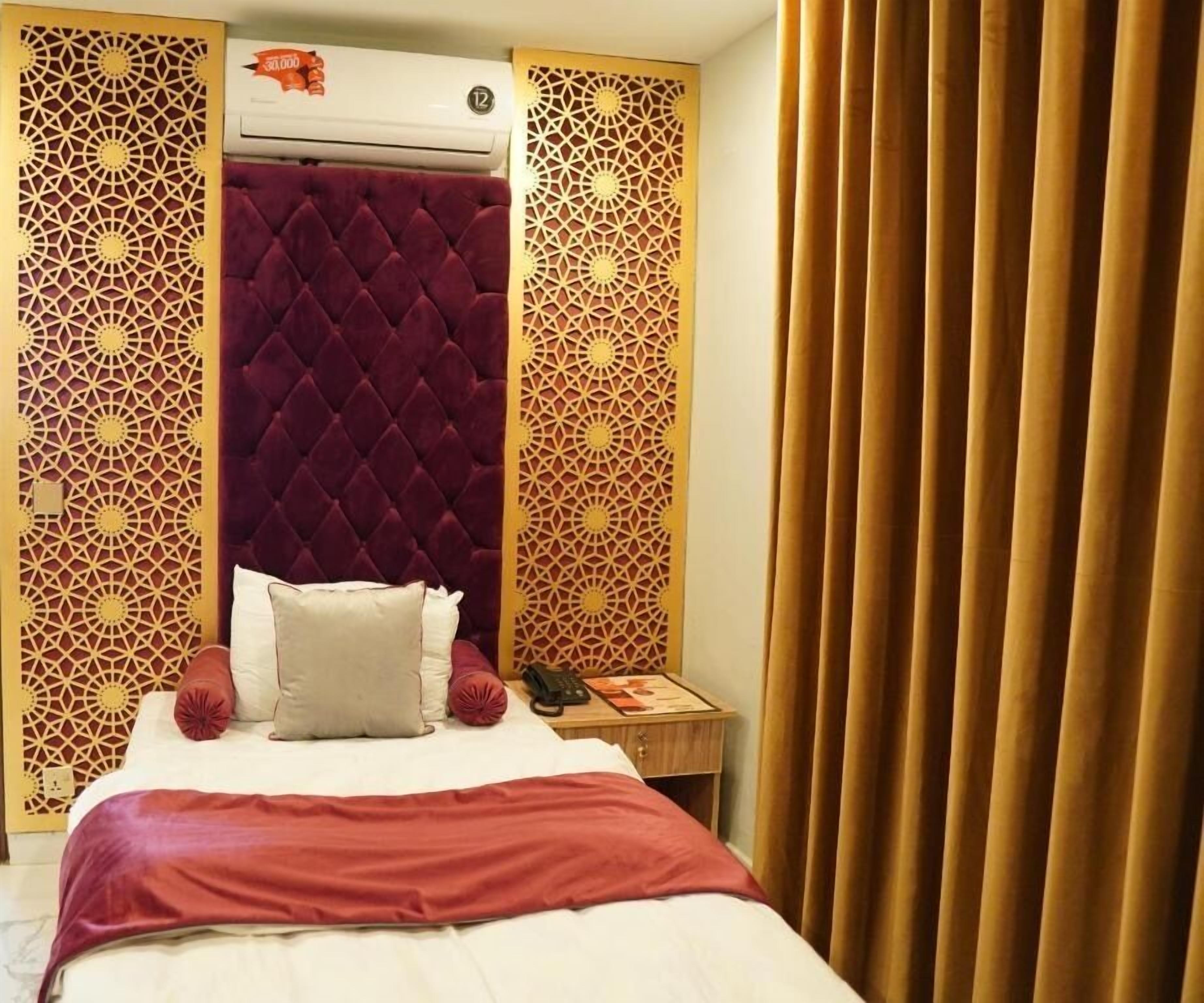 Una habitación de hotel con una cama, cabecera de terciopelo rojo, paneles decorativos, una mesita de noche y una cortina.