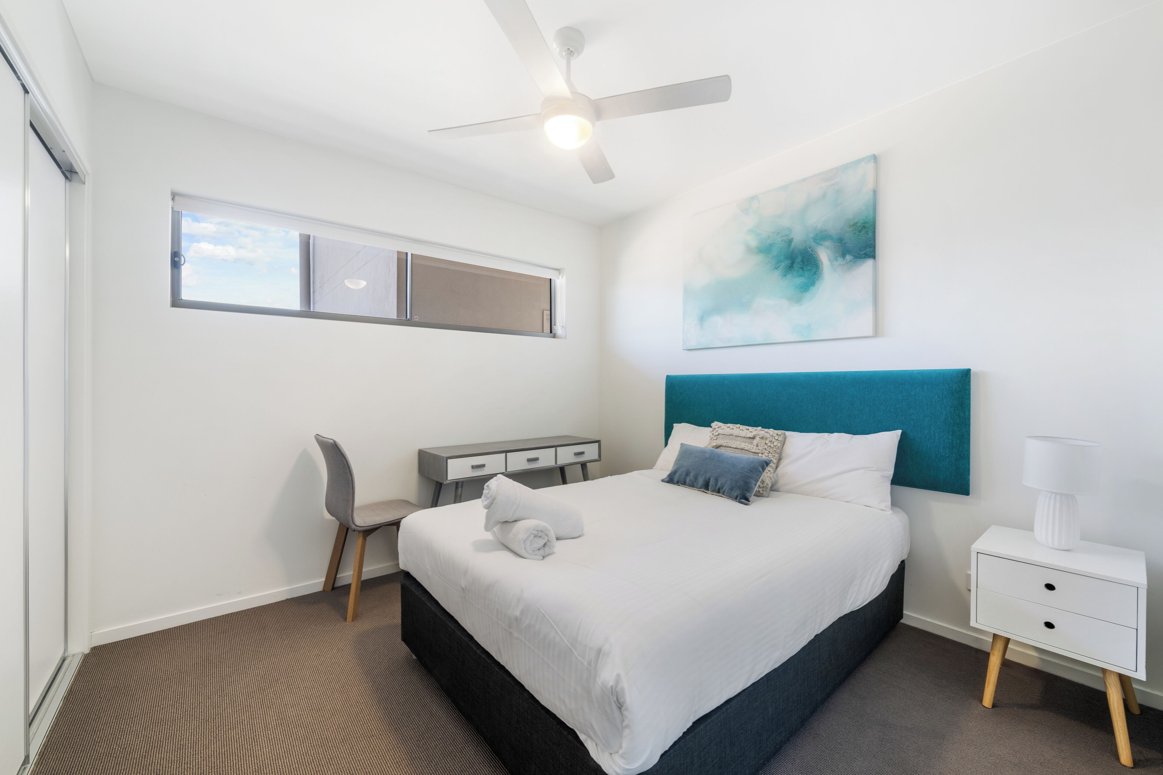 A modern hotel room with a bed, a desk, a chair, a ceiling fan, and a painting on the wall.