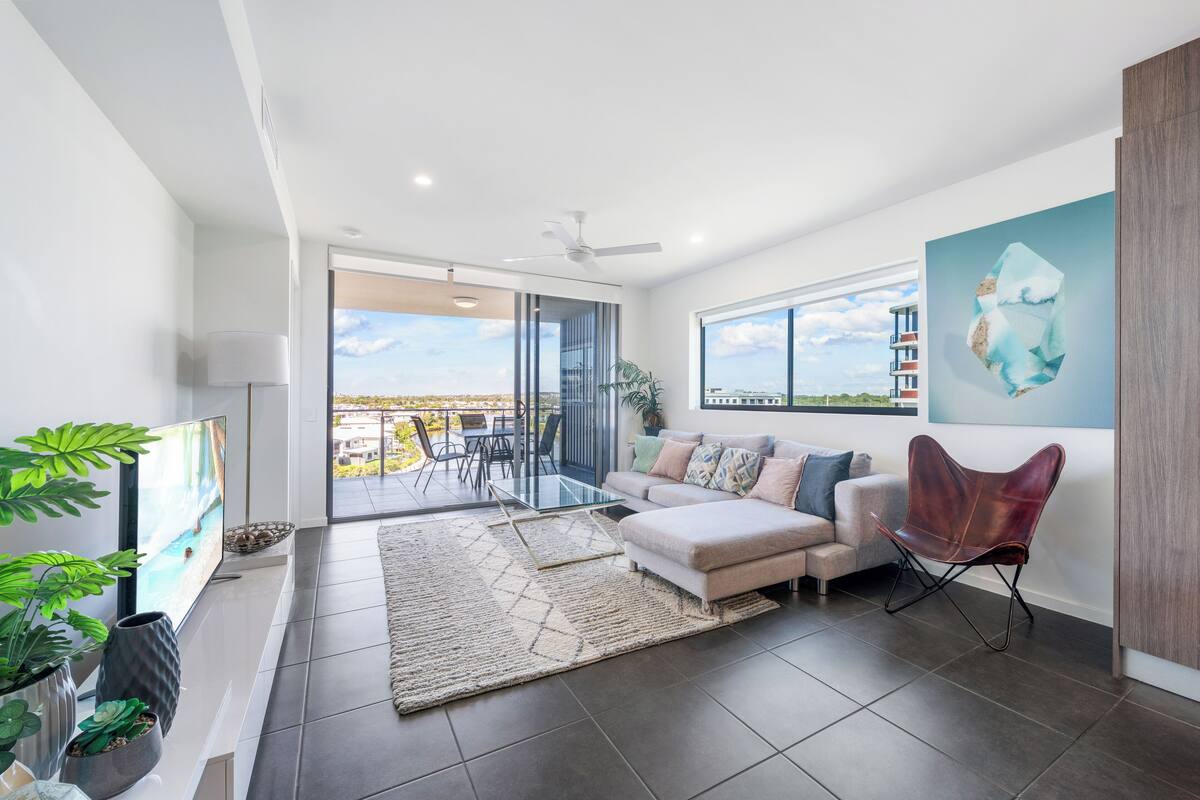 A modern living room with a sofa, a glass coffee table, and a TV. There is a balcony with a view of the city.
