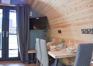 A wooden interior with a dining table set for four, a television mounted on the wall, and a view through a glass door.