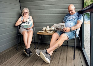 Two people seated on chairs on a wooden deck, with a table between them holding cups and a teapot.
