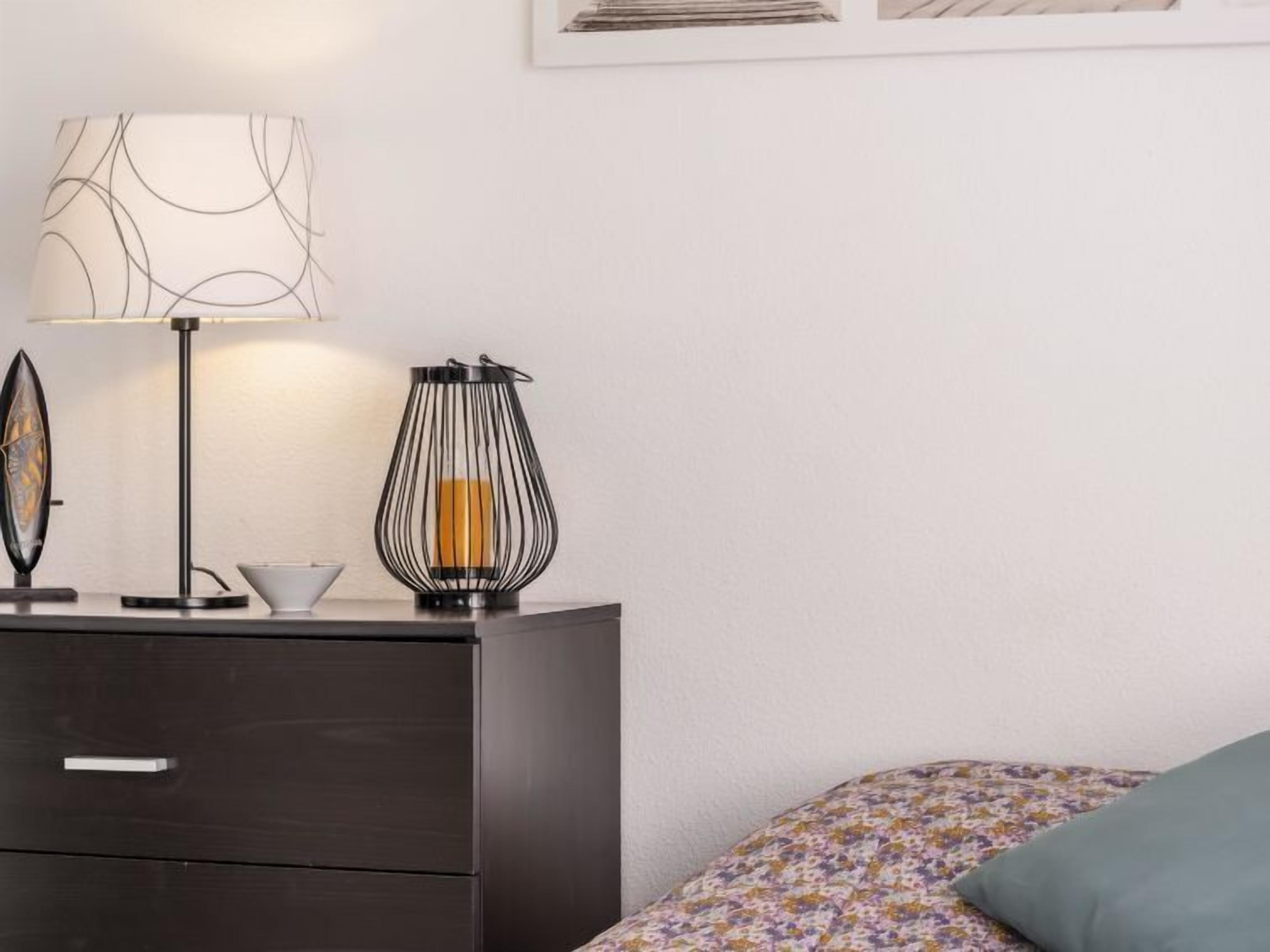 Un dormitorio con una mesita de noche de madera oscura, una lámpara con pantalla estampada, un candelabro y una cama con estampado floral.