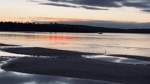 Une plage aux eaux calmes où se reflète le ciel, un bateau au loin et une personne qui se promène le long du rivage.