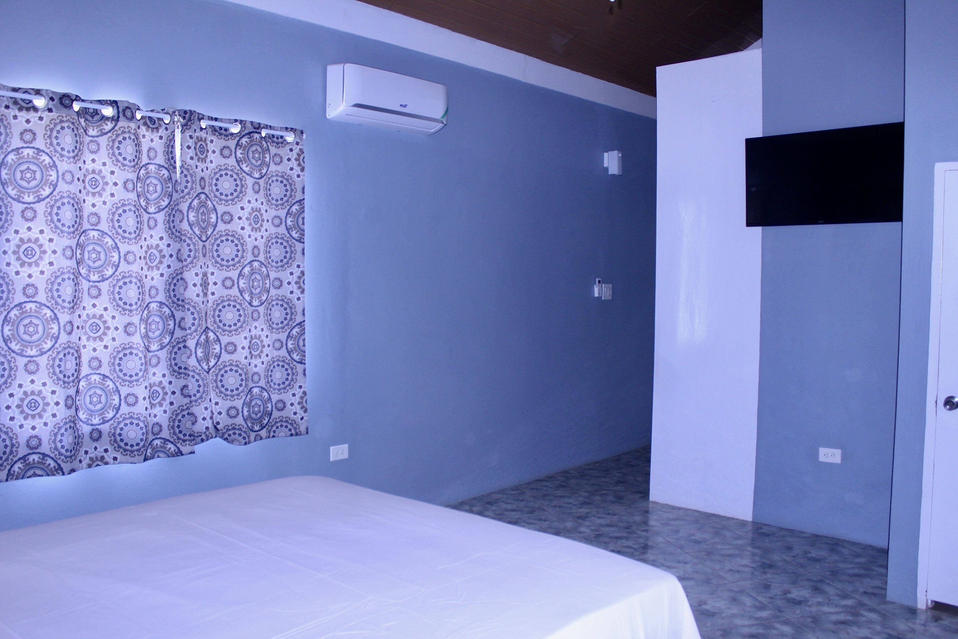 Quarto com cama, cortina estampada, ar condicionado, porta e chão de azulejos.